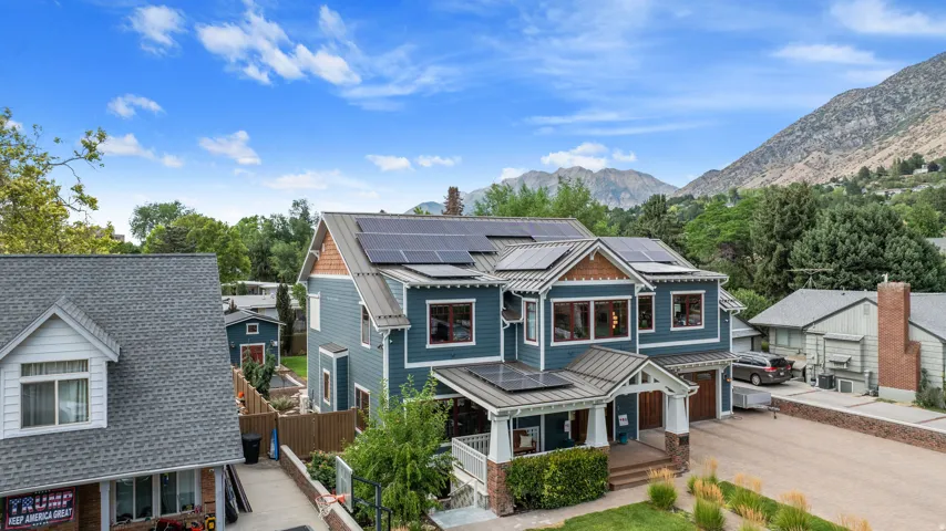 Multi-story residence featuring blue siding, a covered front porch with stone pillars, a two-car garage with wood-finish doors, multiple solar panel arrays, and a mountain backdrop