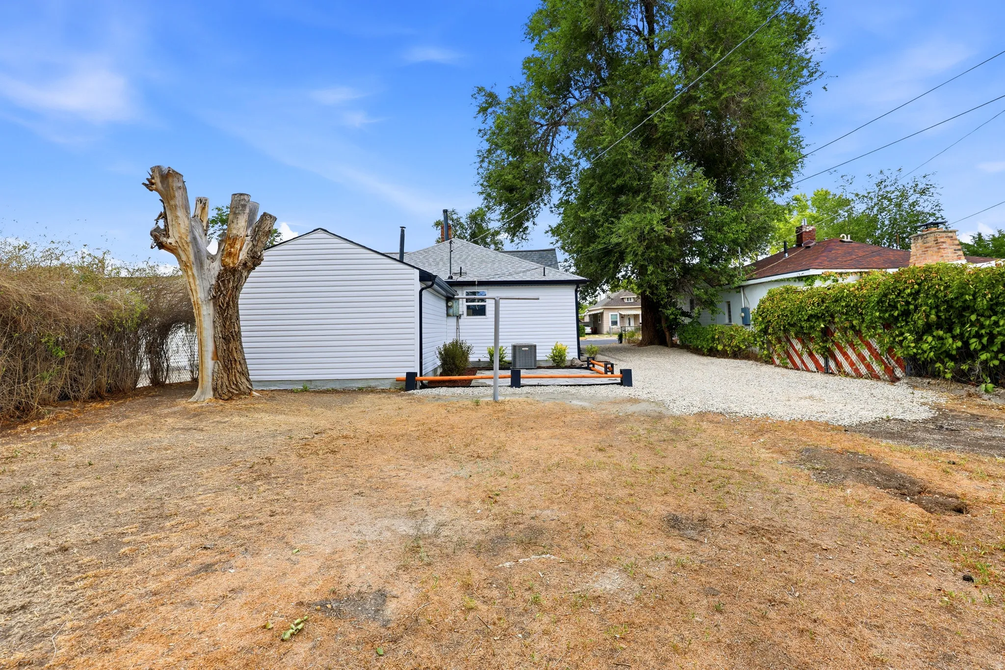 Expansive backyard featuring a white vinyl-sided exterior, a gravel driveway, mature trees, and a wooden patio area