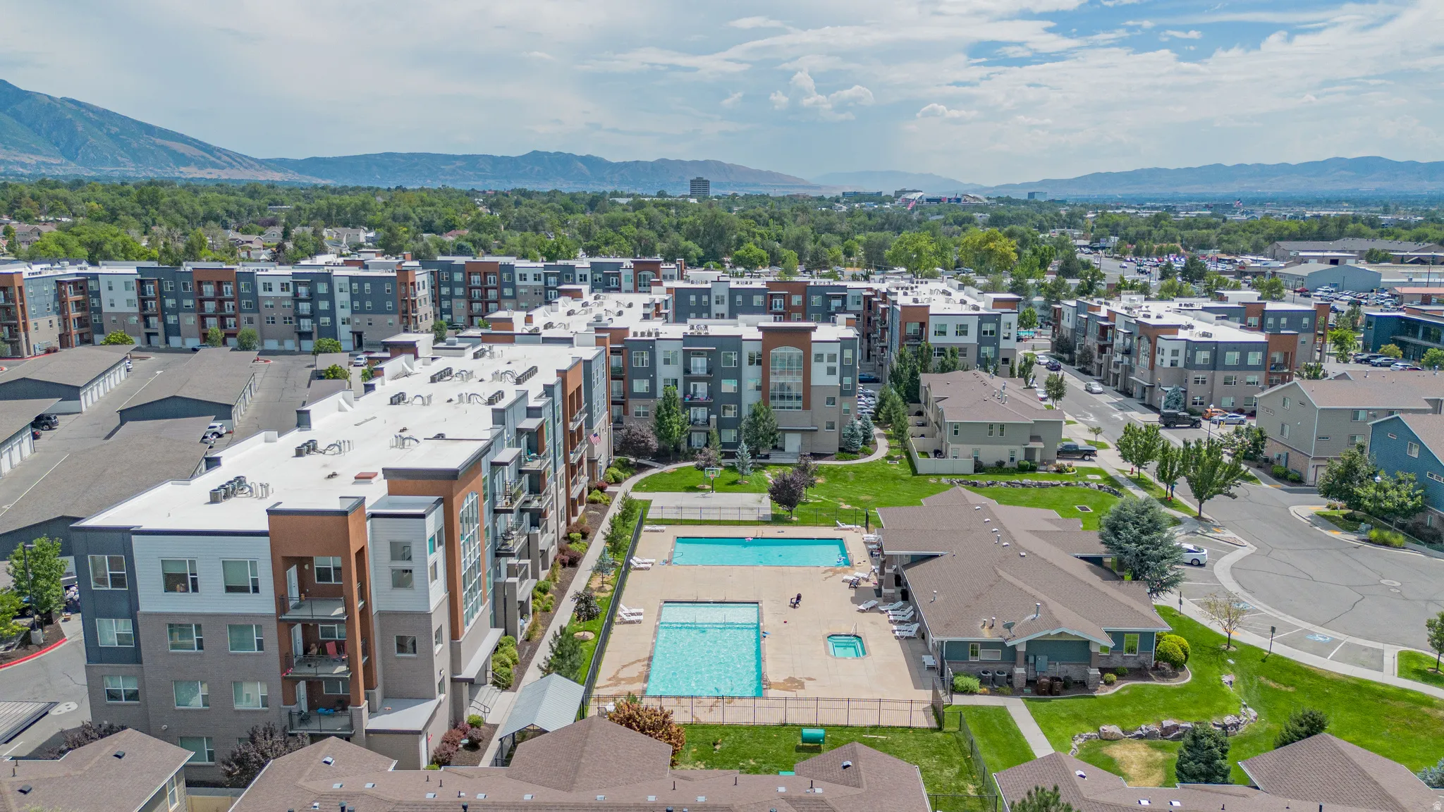 Contemporary multi-story buildings featuring a shared outdoor pool, a separate hot tub, and surrounding green spaces with mature trees