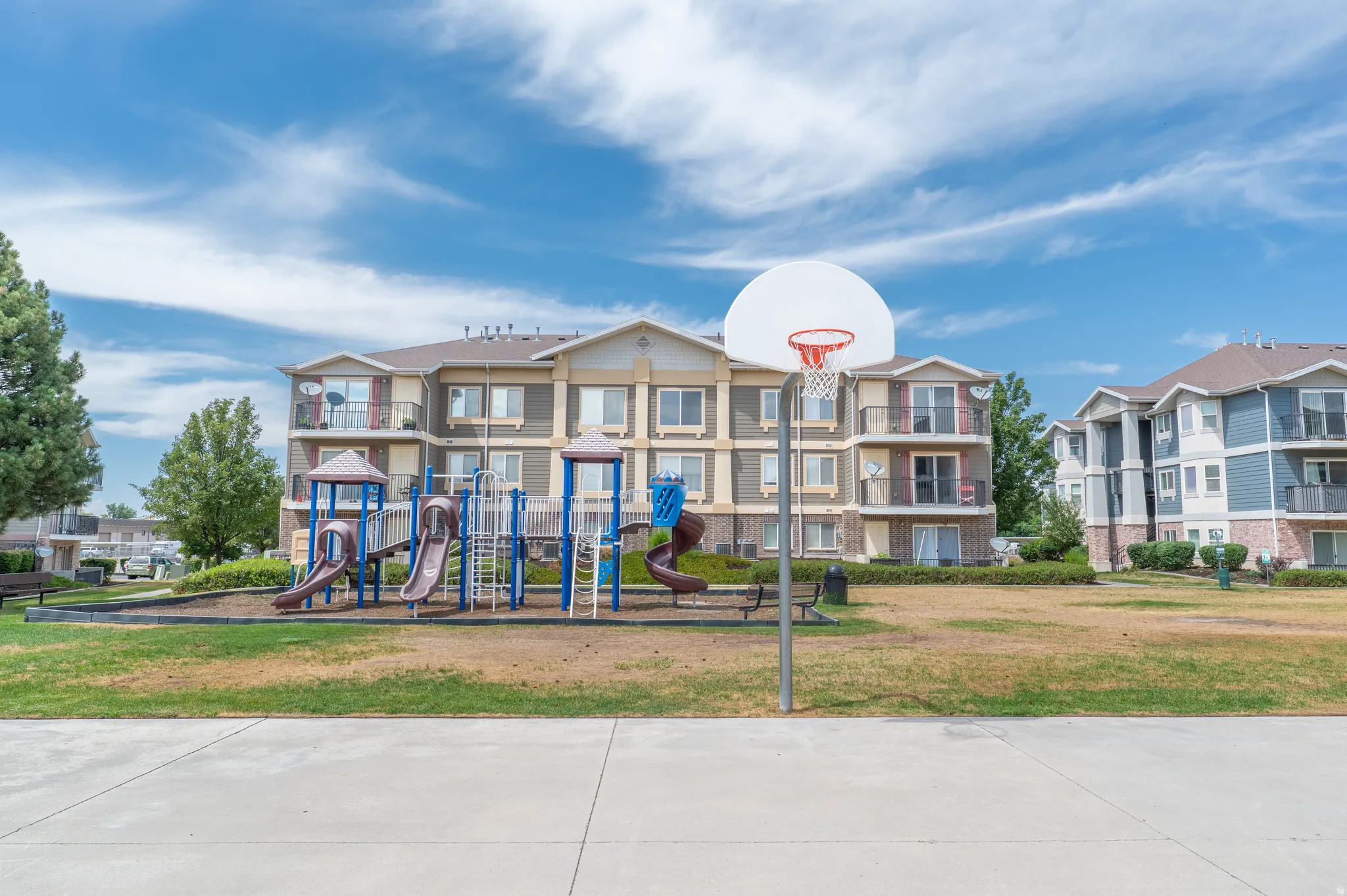 Outdoor recreational area featuring a basketball hoop, a multi-structure playground, and expansive green spaces