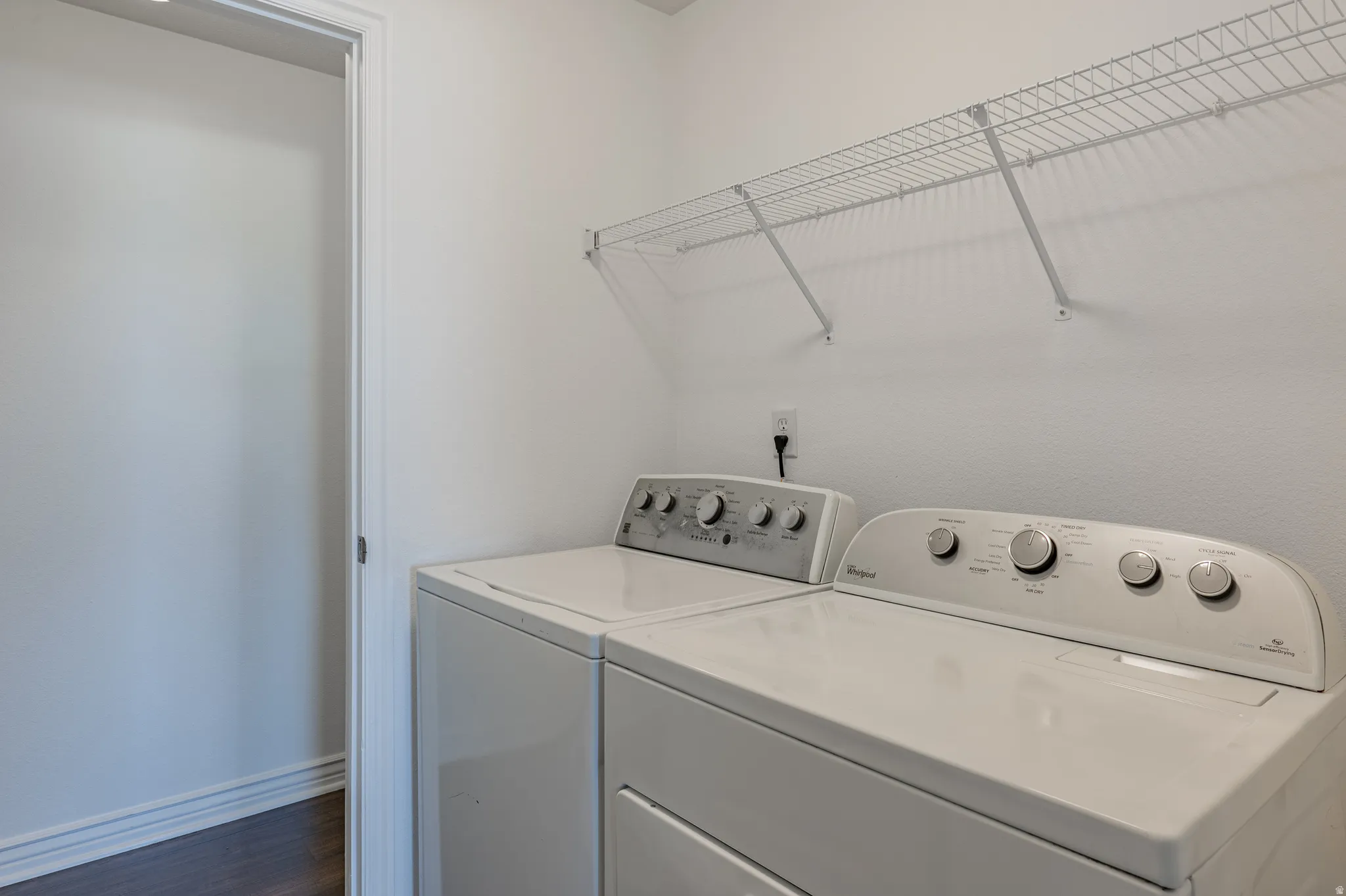 Dedicated laundry area featuring a white wire shelving unit, contrasting dark wood-finish flooring, and a neutral wall color