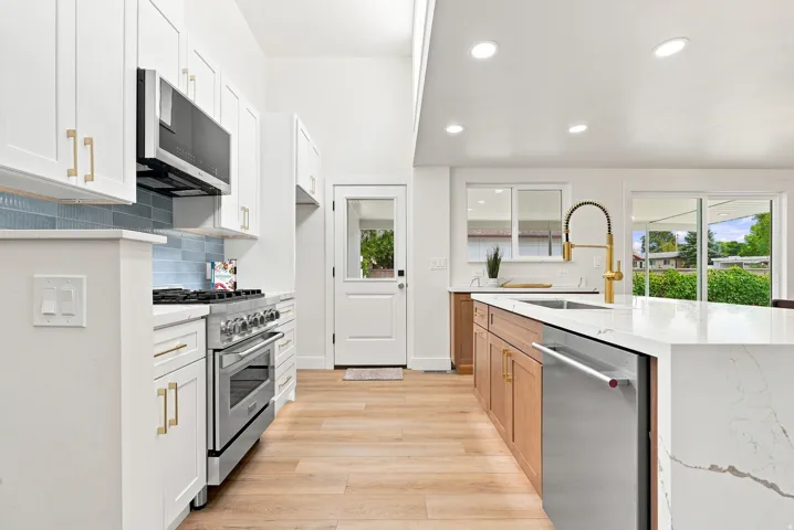 Contemporary kitchen featuring a large island with white stone-finish countertop, wood-finish flooring, white cabinetry with gold-tone hardware, stainless steel appliances, and recessed lighting