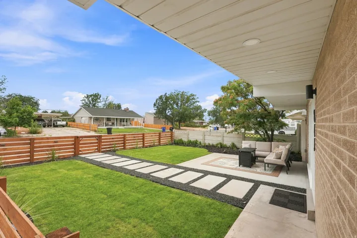 Expansive outdoor living area featuring a covered patio, brick exterior, concrete walkway with dark stone detailing, a manicured lawn, and a slatted wood fence