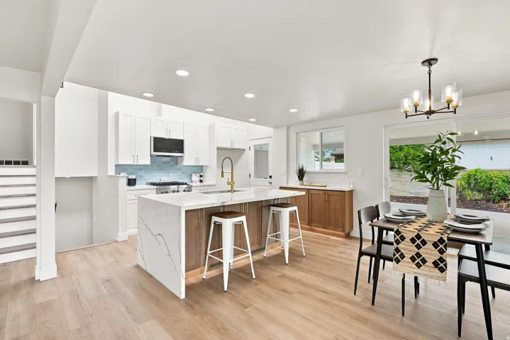 Open-concept living space featuring a modern kitchen with white cabinetry, a waterfall-edge island with seating, a blue tile backsplash, and light wood-finish flooring
