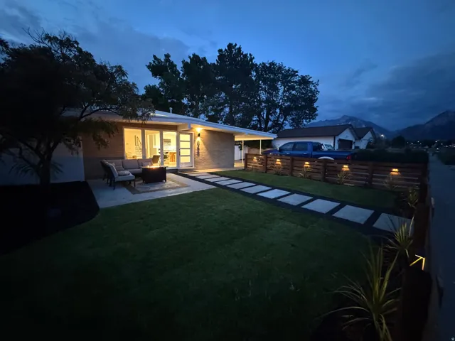 Front exterior featuring a modern brick facade, concrete patio, concrete stepping stones, manicured lawn, and a wooden privacy fence with integrated lighting