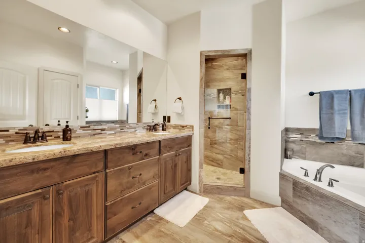 Spacious bathroom featuring a dual vanity with wood-finish cabinetry, a large built-in mirror, a glass-enclosed shower with tiled surround, a corner soaking tub, and coordinating tile flooring