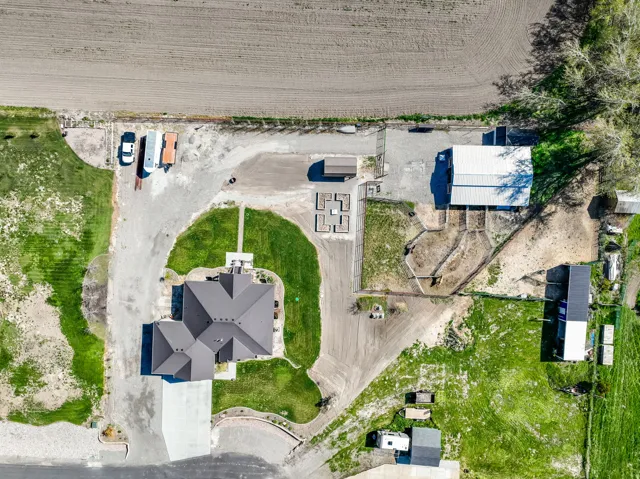 Expansive property featuring a contemporary residence with a dark shingle roof, a substantial detached outbuilding with a light-colored roof, multiple gravel driveways, an established lawn area around the main residence, and several enclosed garden or pen