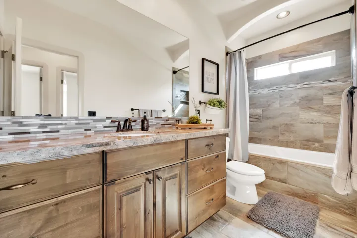 Bathroom featuring a wood-finish double vanity with a light-toned countertop, mosaic tile backsplash, and bronze-finish fixtures