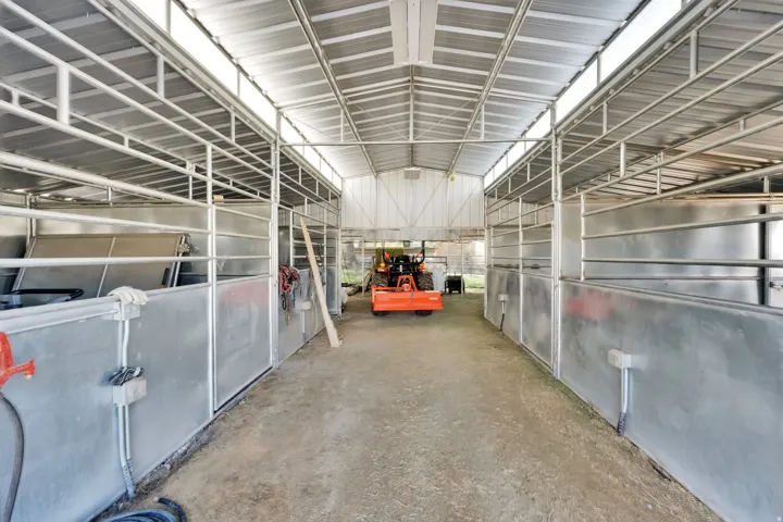 Metal-framed outbuilding featuring multiple stalls with metal gates and lower panels, a high ceiling with exposed metal trusses, and translucent roof panels providing natural light