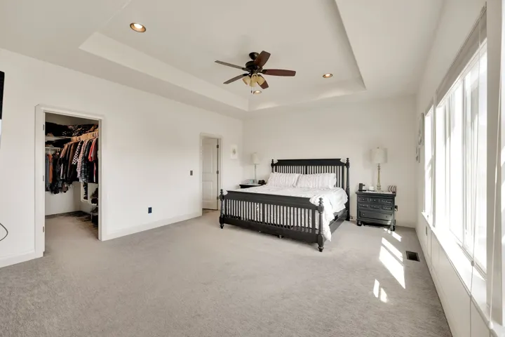 Spacious bedroom featuring light-colored carpeting, a tray ceiling with recessed lighting and a ceiling fan, and multiple windows with white trim