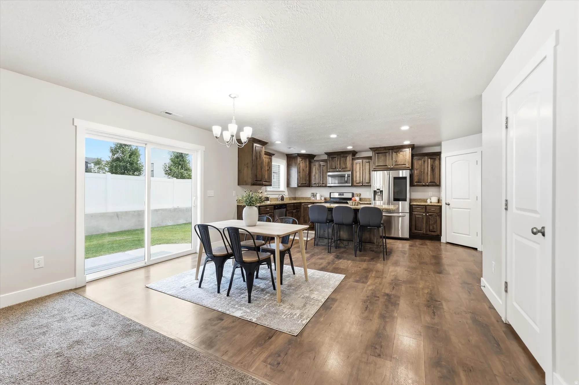Open-concept living space featuring wood-finish flooring, a kitchen with dark cabinetry and stainless steel appliances, a dining area with a chandelier, and sliding glass doors to the backyard