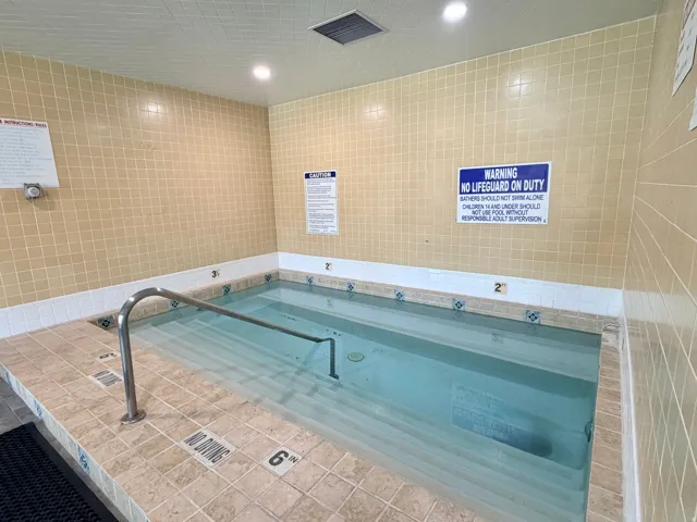 Indoor pool with integrated steps and a stainless steel handrail, surrounded by tan square wall tiles and contrasting decorative border tiles
