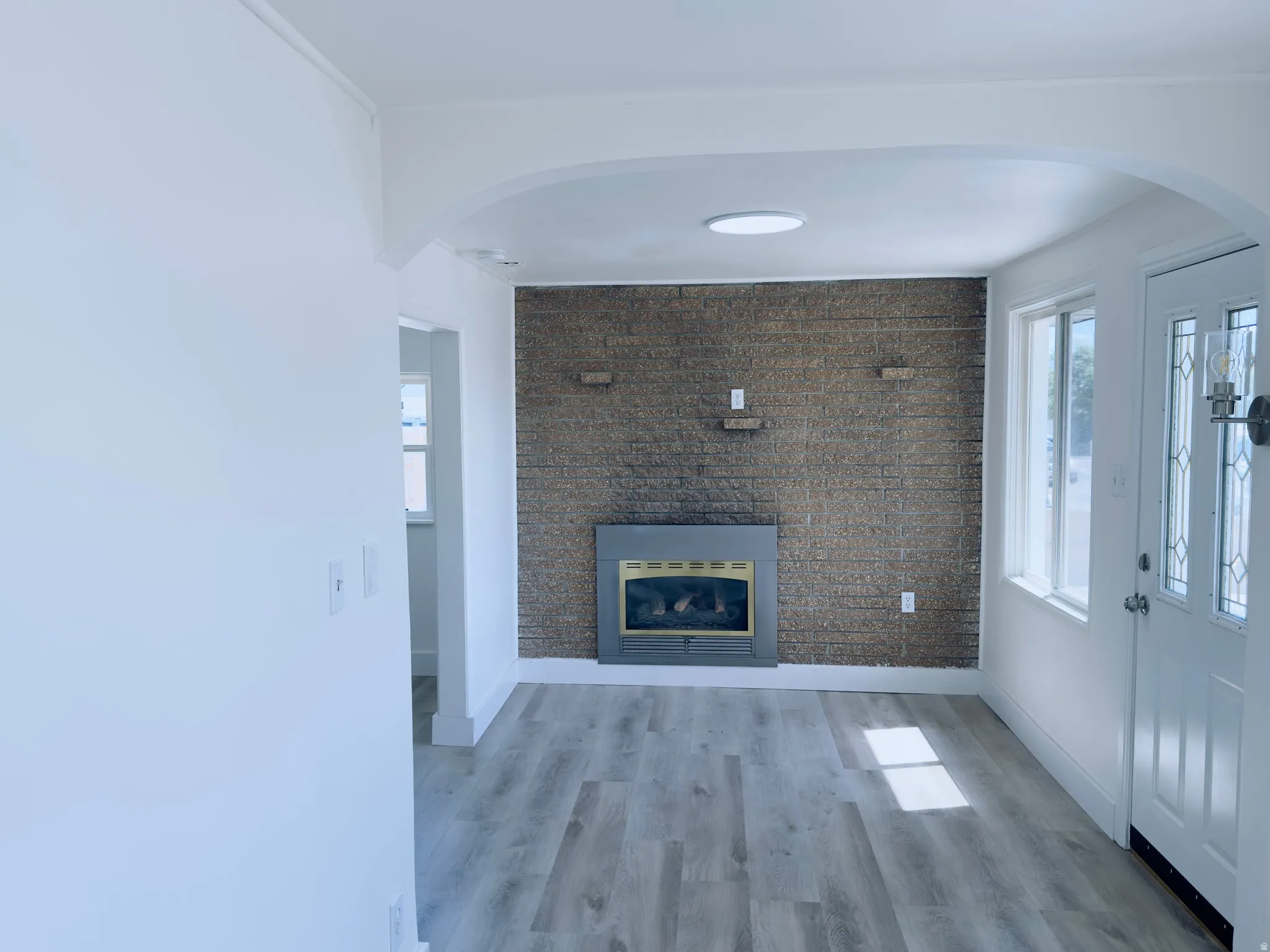 Living area featuring a textured accent wall with an inset fireplace, light wood-finish flooring, a decorative front entry door with leaded glass, and an arched entryway