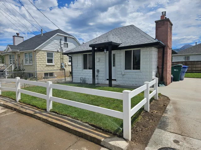 Single-story residence featuring white textured exterior siding, a dark shingle roof, a red brick chimney, and a white split-rail fence enclosing a grass lawn