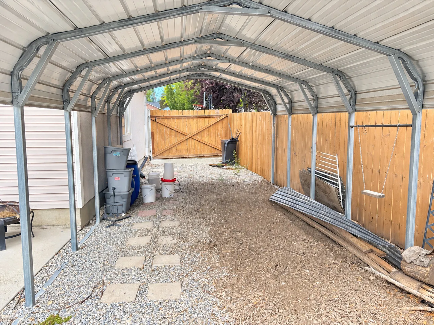 Metal carport structure with a corrugated roof, gravel ground cover, stepping stones, and a wood privacy fence