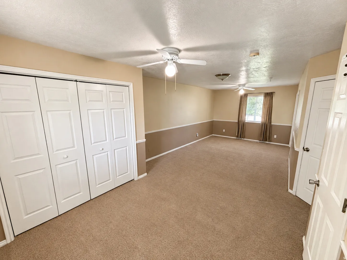 Spacious bedroom featuring neutral wall tones with contrasting chair rail detail, plush carpet flooring, ceiling fan with integrated lighting, and dual closet access including a multi-panel sliding door system