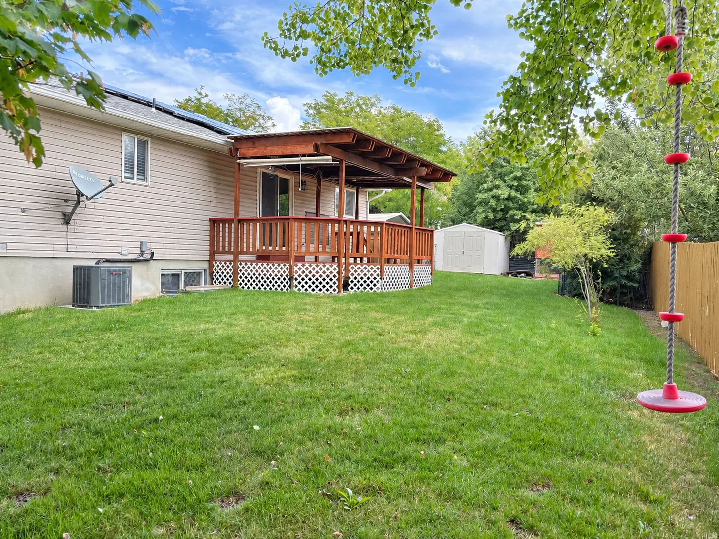 Expansive green lawn featuring a covered deck, white lattice skirting, and a storage shed