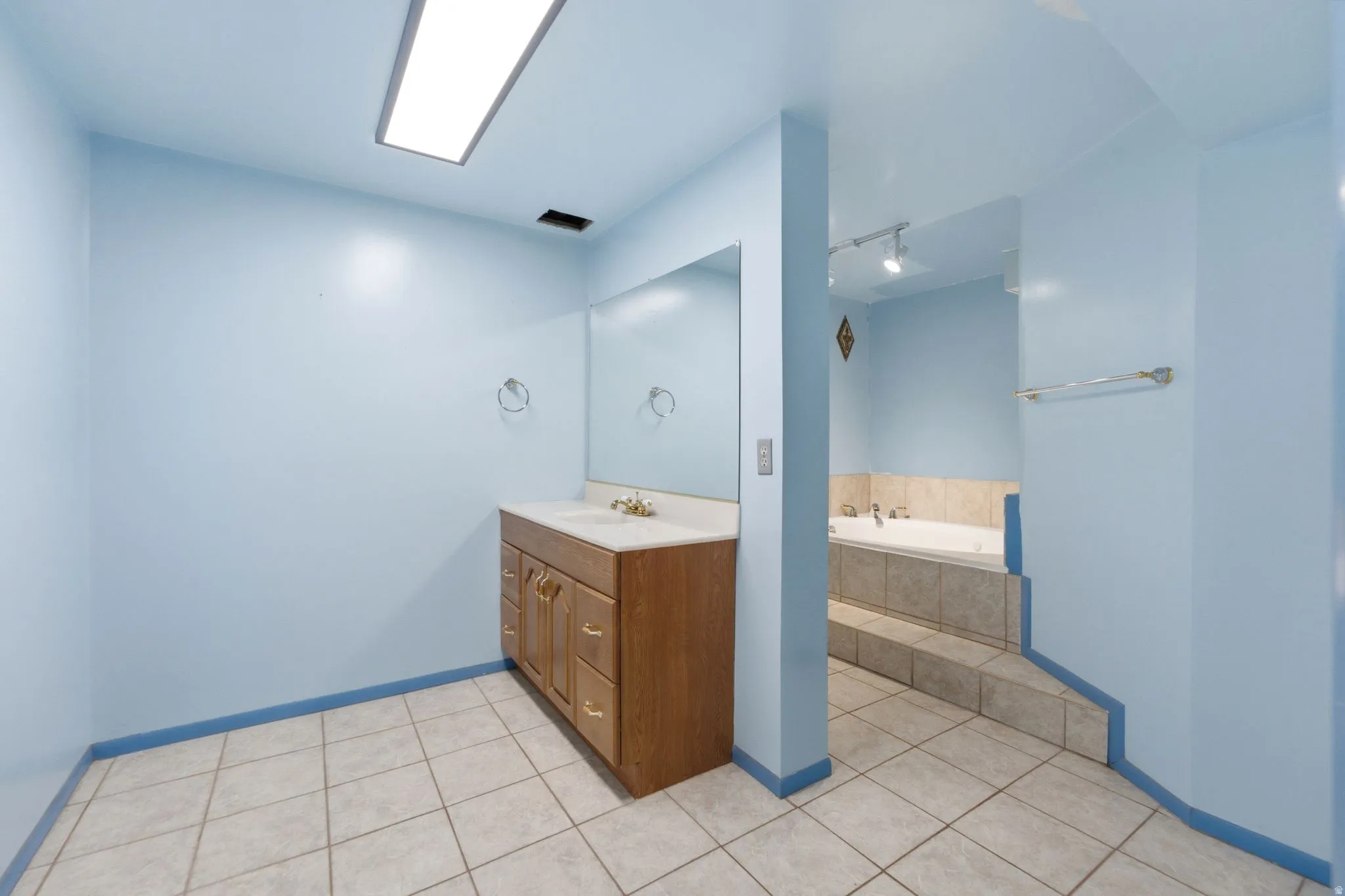 Bathroom featuring a wood vanity with countertop sink, large wall-mounted mirror, overhead fluorescent lighting, ceramic tile flooring, and an oversized tub.