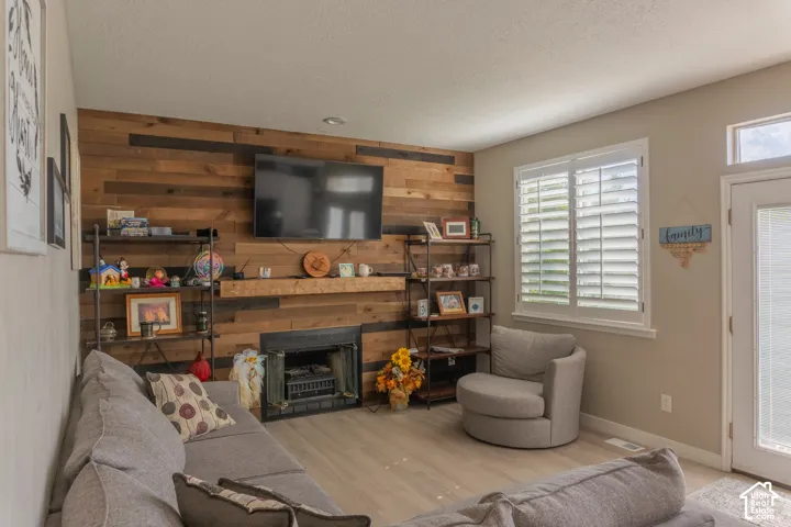 Living area featuring a floor-to-ceiling wood plank accent wall, a built-in fireplace with a wood mantle, light-toned wood-finish flooring, and a large window with plantation shutters