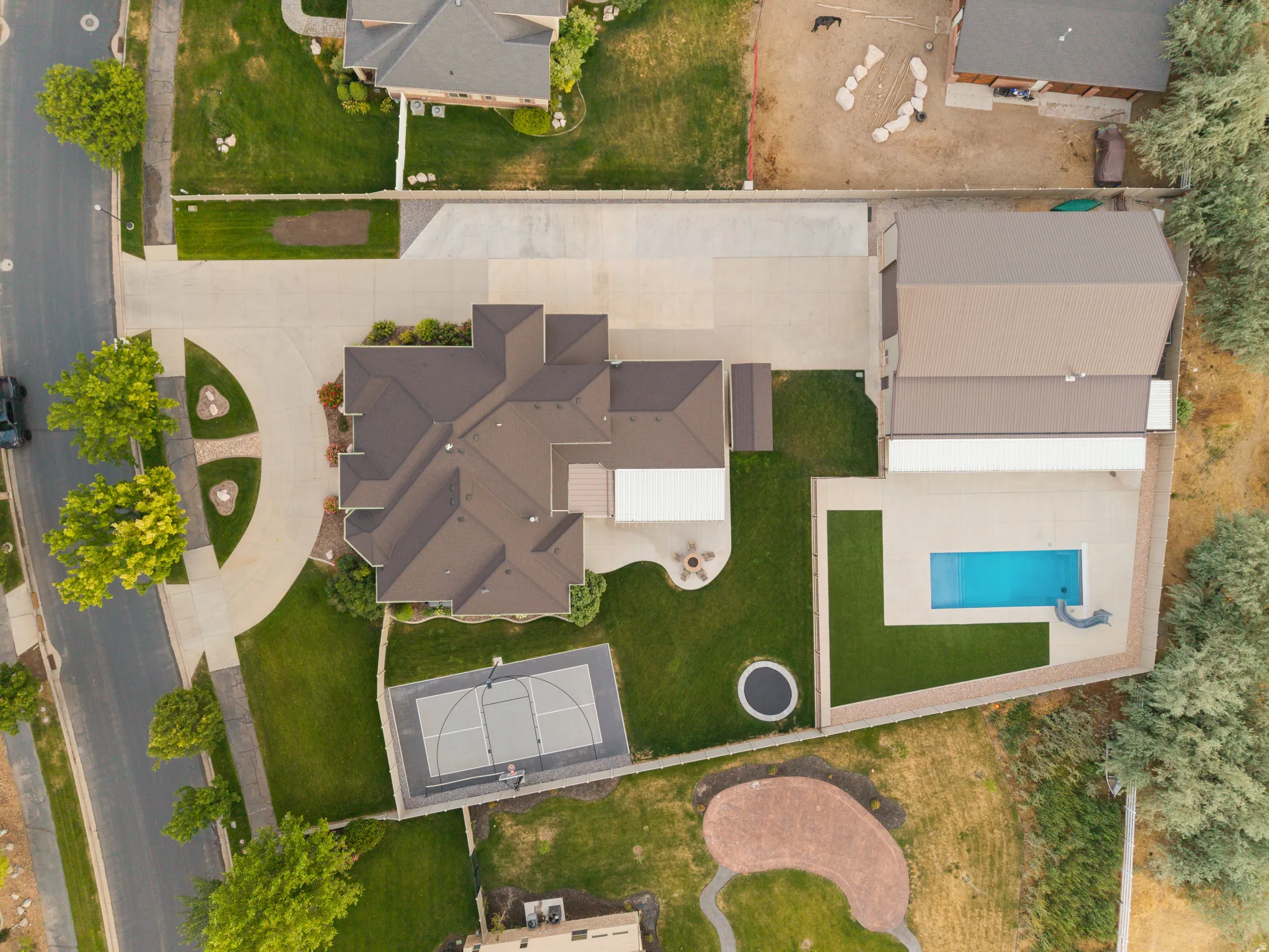 Expansive property featuring a multi-level home with a dark shingle roof, a large concrete driveway, a dedicated sport court, an in-ground swimming pool with a slide, and a separate outbuilding with a metal roof