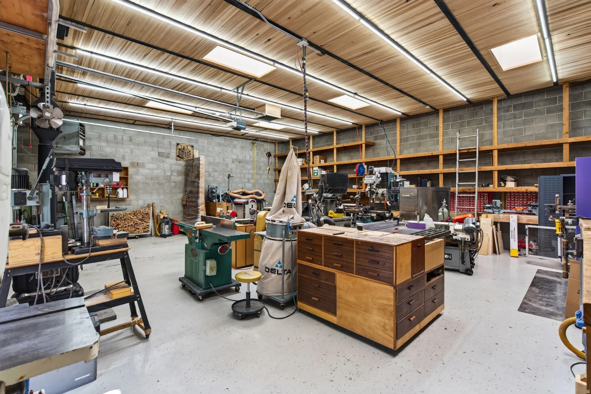 Spacious workshop featuring a wood-paneled ceiling with integrated fluorescent lighting and skylights, concrete block walls, and light-colored epoxy flooring