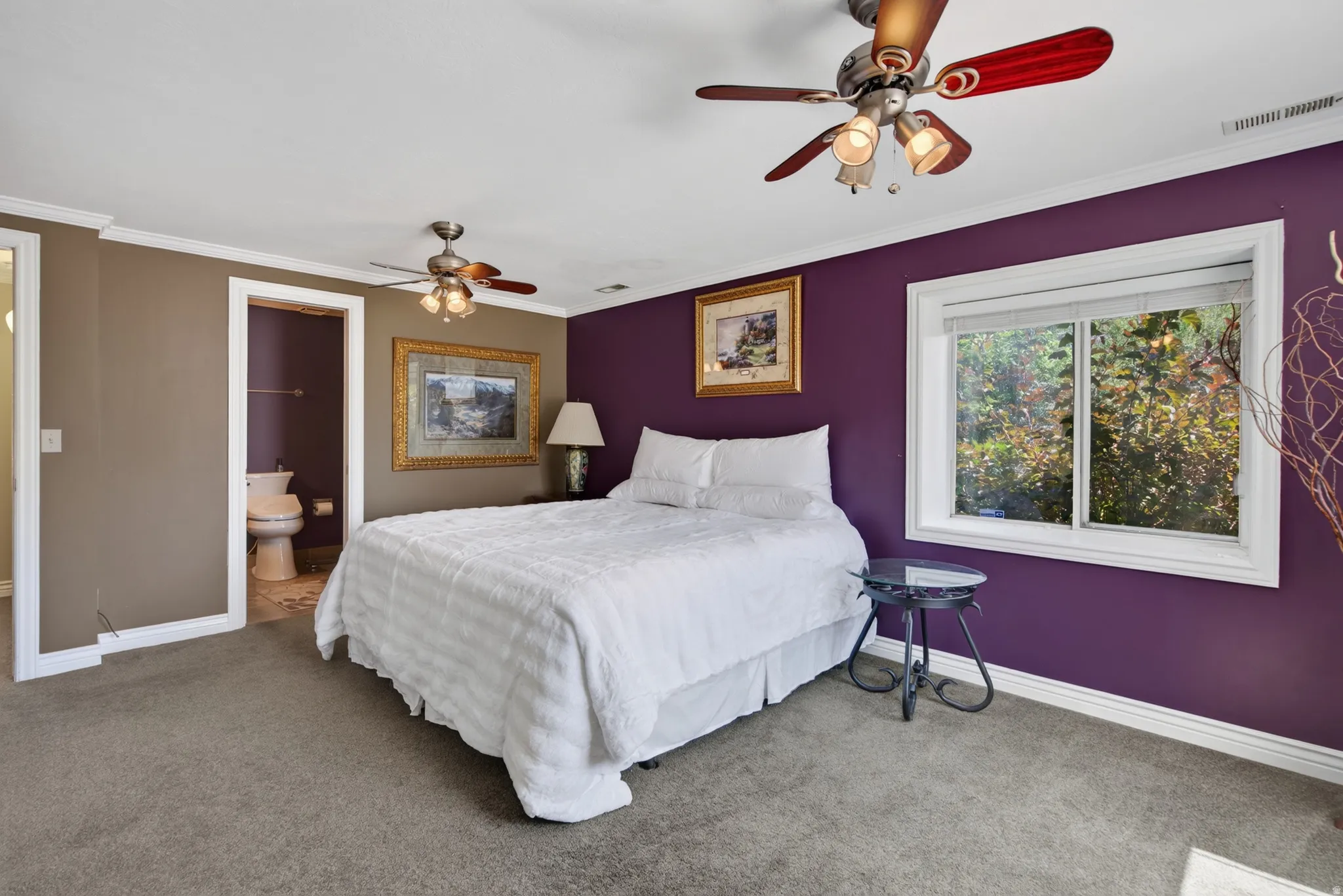 Bedroom featuring a large window, two ceiling fans with light fixtures, and a private attached bathroom with a toilet and wall-mounted towel bar
