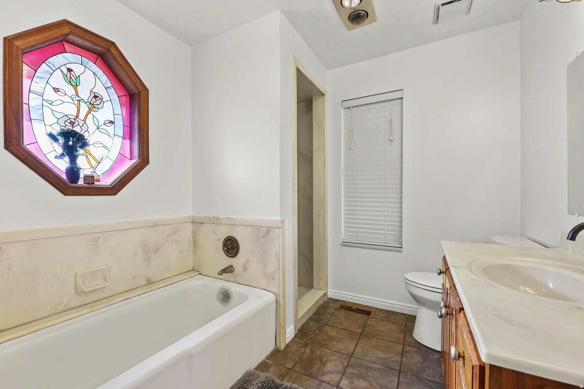 Bathroom featuring a built-in bathtub with a tile surround, an octagonal stained-glass window, a single vanity with an integrated sink and wood-finish cabinetry, and tiled flooring