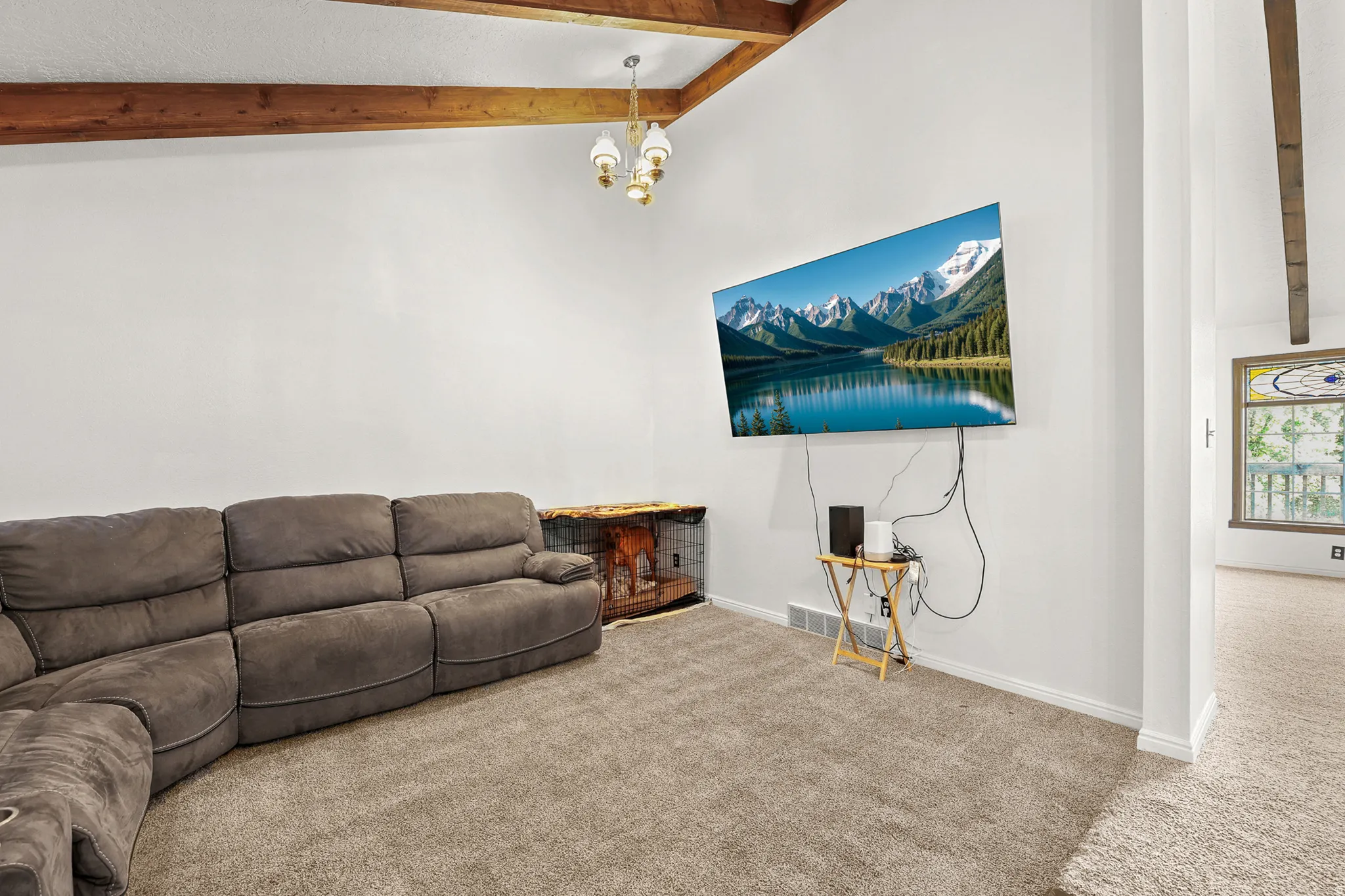Vaulted ceiling with exposed wood beams, overhead chandelier, wall-mounted television, and light-toned carpeting