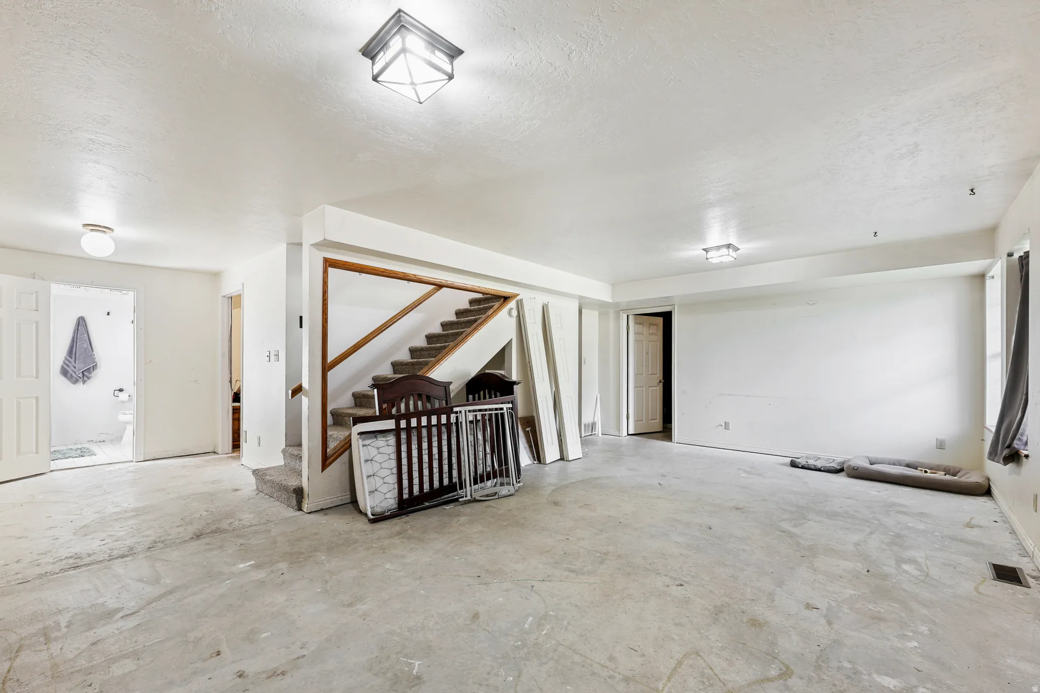 Spacious lower-level room featuring an exposed concrete floor, white walls, and a staircase with wood railing