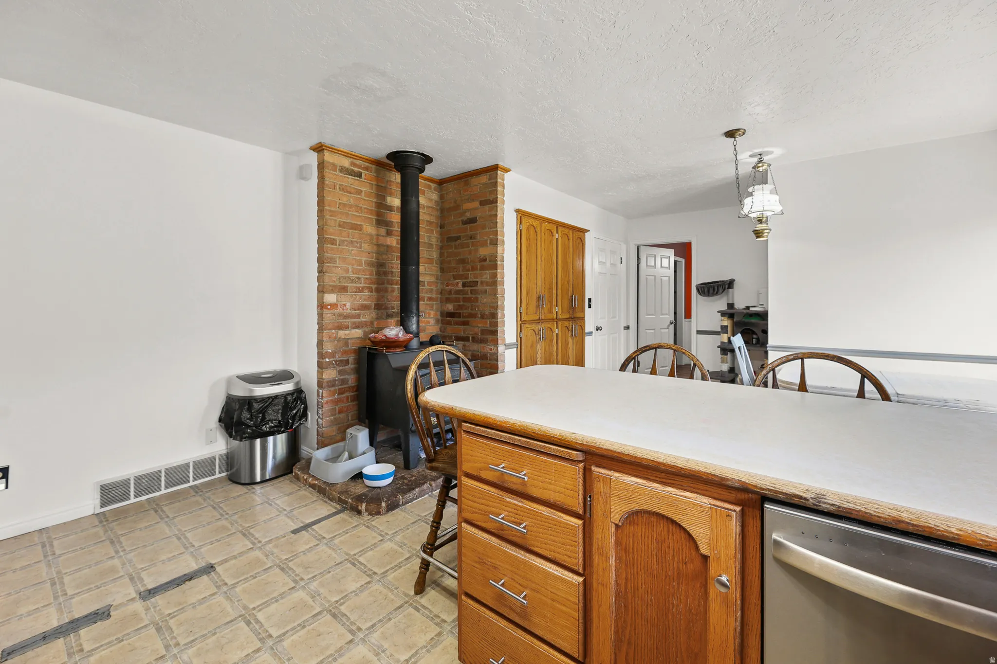 Kitchen area featuring a brick accent wall with a wood-burning stove, light-toned countertop, wood-finish cabinetry with silver hardware, an integrated dishwasher, and patterned tile flooring