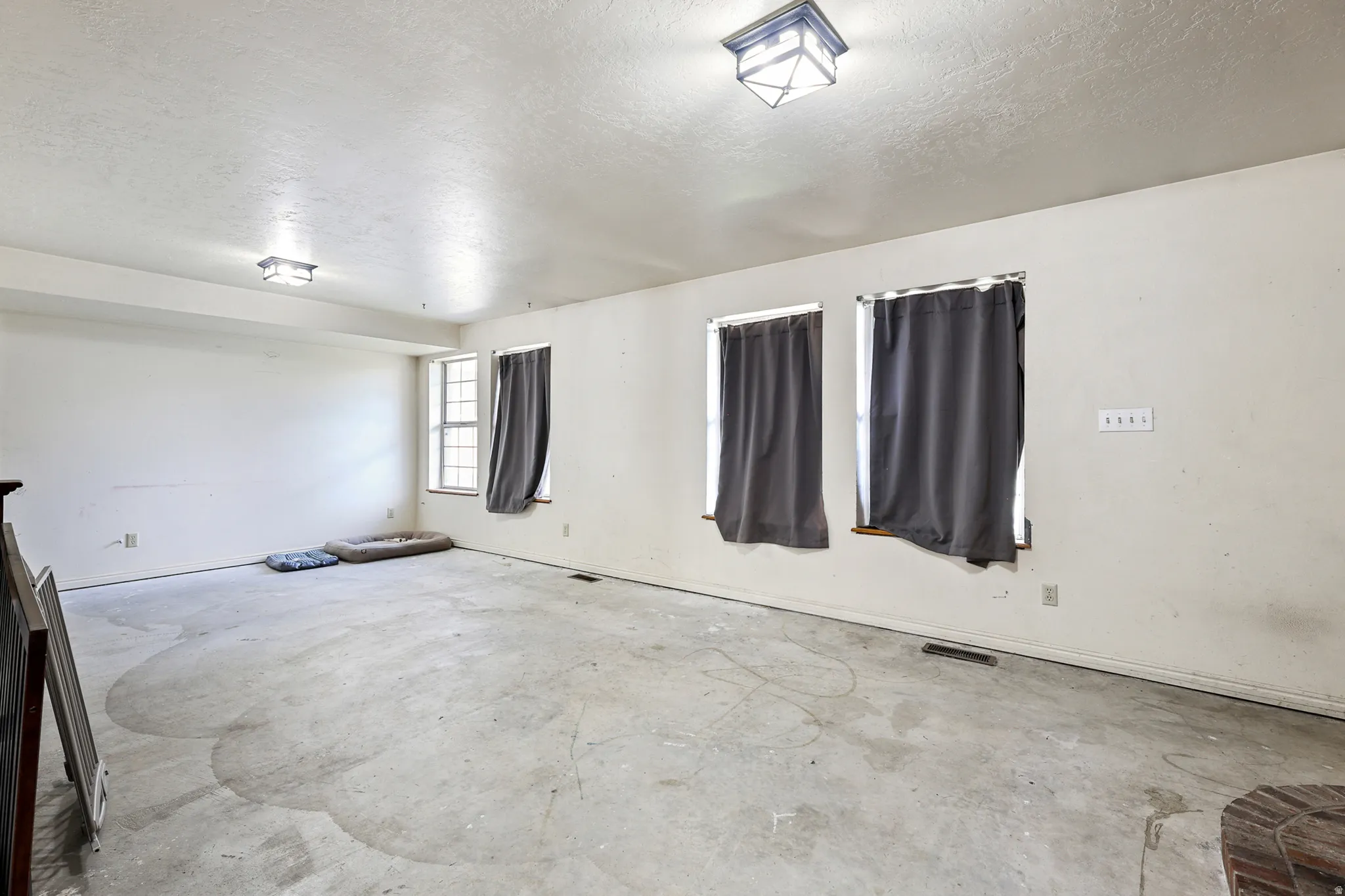 Spacious room featuring three windows, two ceiling-mounted light fixtures, and raw concrete flooring