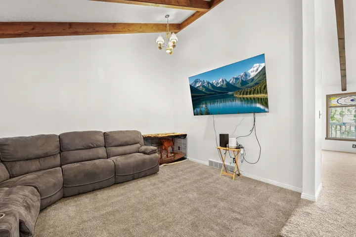 Vaulted ceiling with exposed wood beams, overhead chandelier, wall-mounted television, and light-toned carpeting