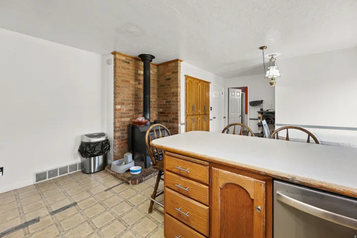 Kitchen area featuring a brick accent wall with a wood-burning stove, light-toned countertop, wood-finish cabinetry with silver hardware, an integrated dishwasher, and patterned tile flooring