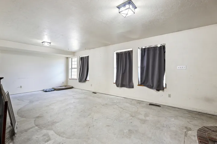 Spacious room featuring three windows, two ceiling-mounted light fixtures, and raw concrete flooring