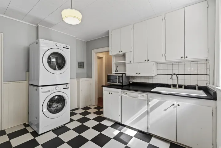 Kitchen area featuring stacked white washer and dryer, checkerboard flooring, white cabinetry, dark countertops, and a white tiled backsplash