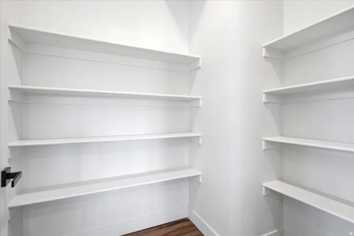Walk-in pantry featuring multiple levels of white shelving, dark wood-finish flooring, and a modern black door handle