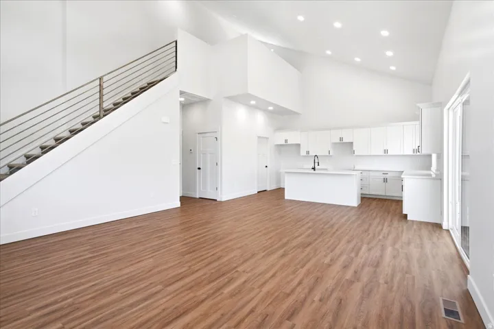 Open-concept living space featuring wood-finish flooring, a contemporary kitchen with white cabinetry and an island, a modern staircase with metal railing, vaulted ceilings, and recessed lighting