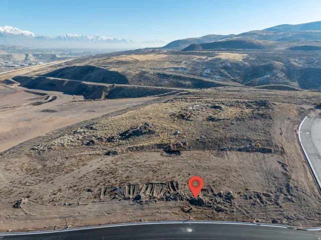 Expansive undeveloped lot featuring rugged terrain, a newly paved road bordering the property, and distant snow-capped mountains under a clear sky