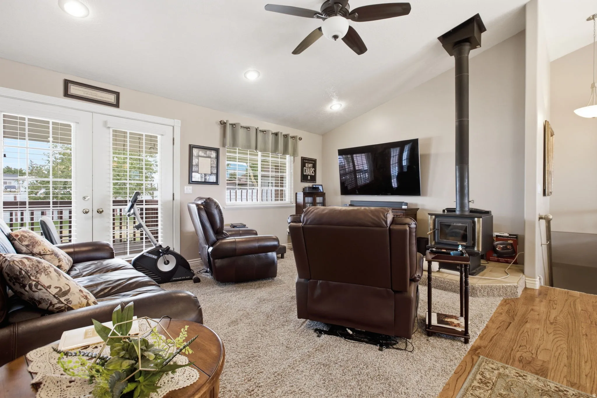 Open concept living area featuring vaulted ceilings, recessed lighting, a ceiling fan, a wood-burning stove with a hearth, and double French doors opening to an exterior deck