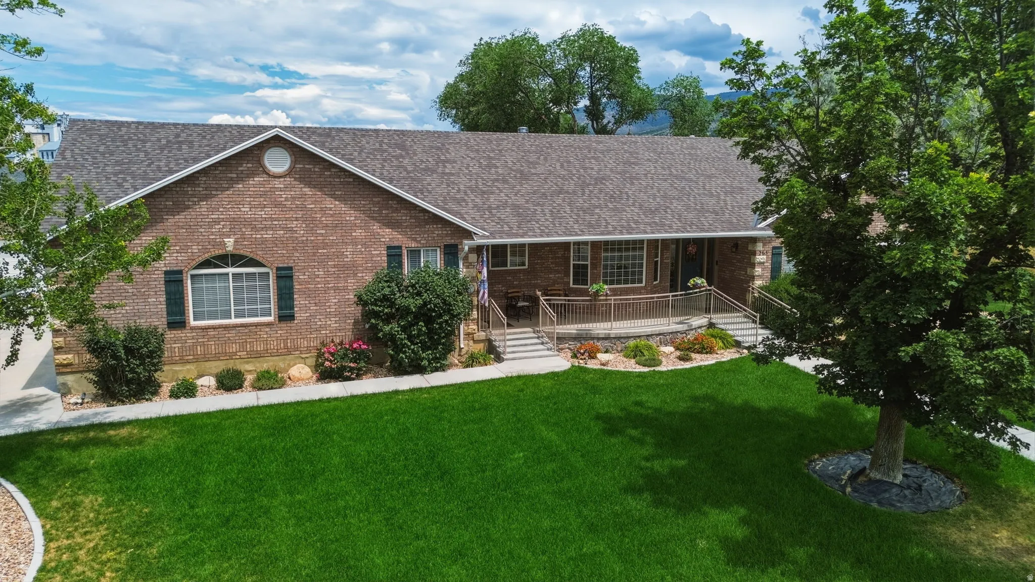Brick façade residence featuring a covered porch with railings, mature front yard trees, and a well-maintained lawn