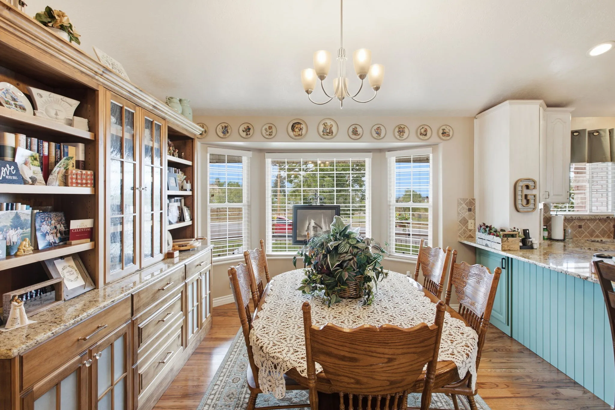 Bright dining area featuring wood-finish flooring, a contemporary chandelier, a built-in wooden hutch with glass-front cabinetry, a bay window with multiple panes, and a partial view of a kitchen island with a light blue beadboard base and granite counter
