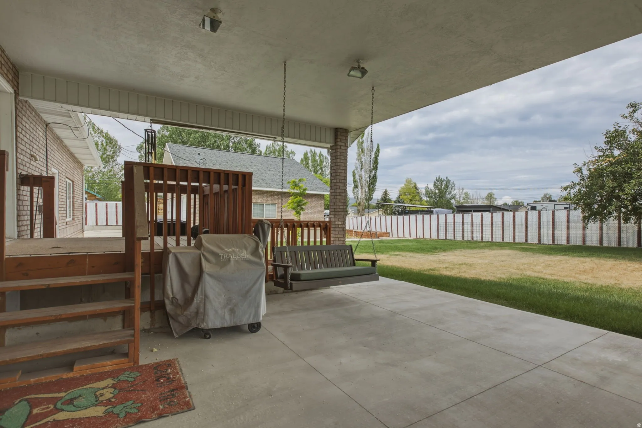 Covered concrete patio extending to a grass yard, featuring a raised wood deck with balusters and stairs