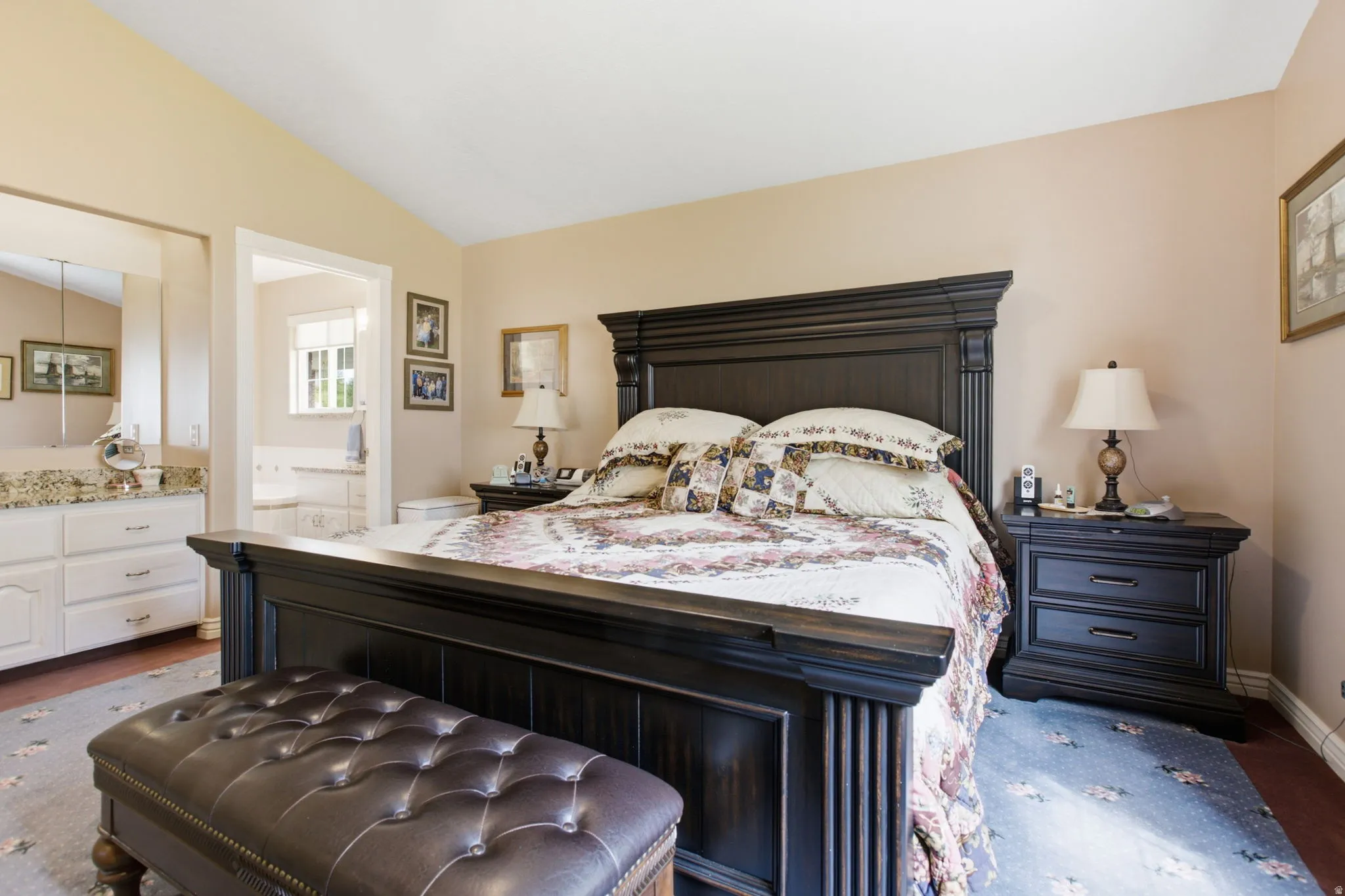 Vaulted ceiling bedroom with wood-finish flooring, an open doorway to a bathroom with a dual vanity, and a window providing natural light