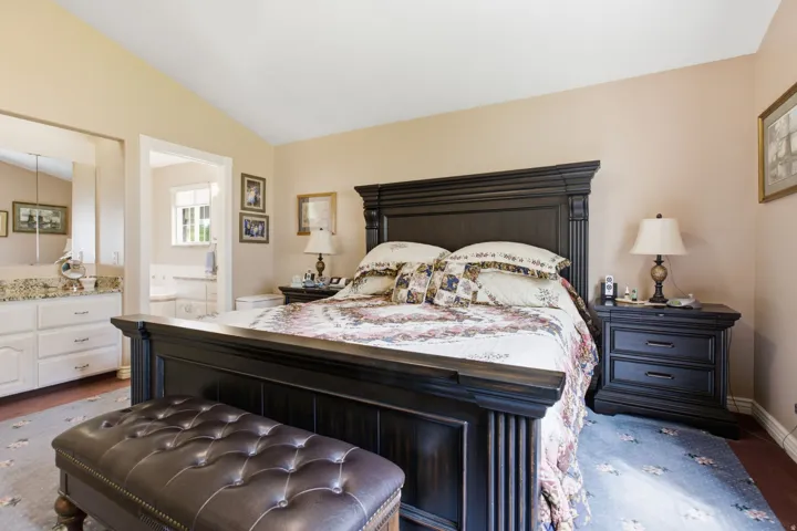 Vaulted ceiling bedroom with wood-finish flooring, an open doorway to a bathroom with a dual vanity, and a window providing natural light