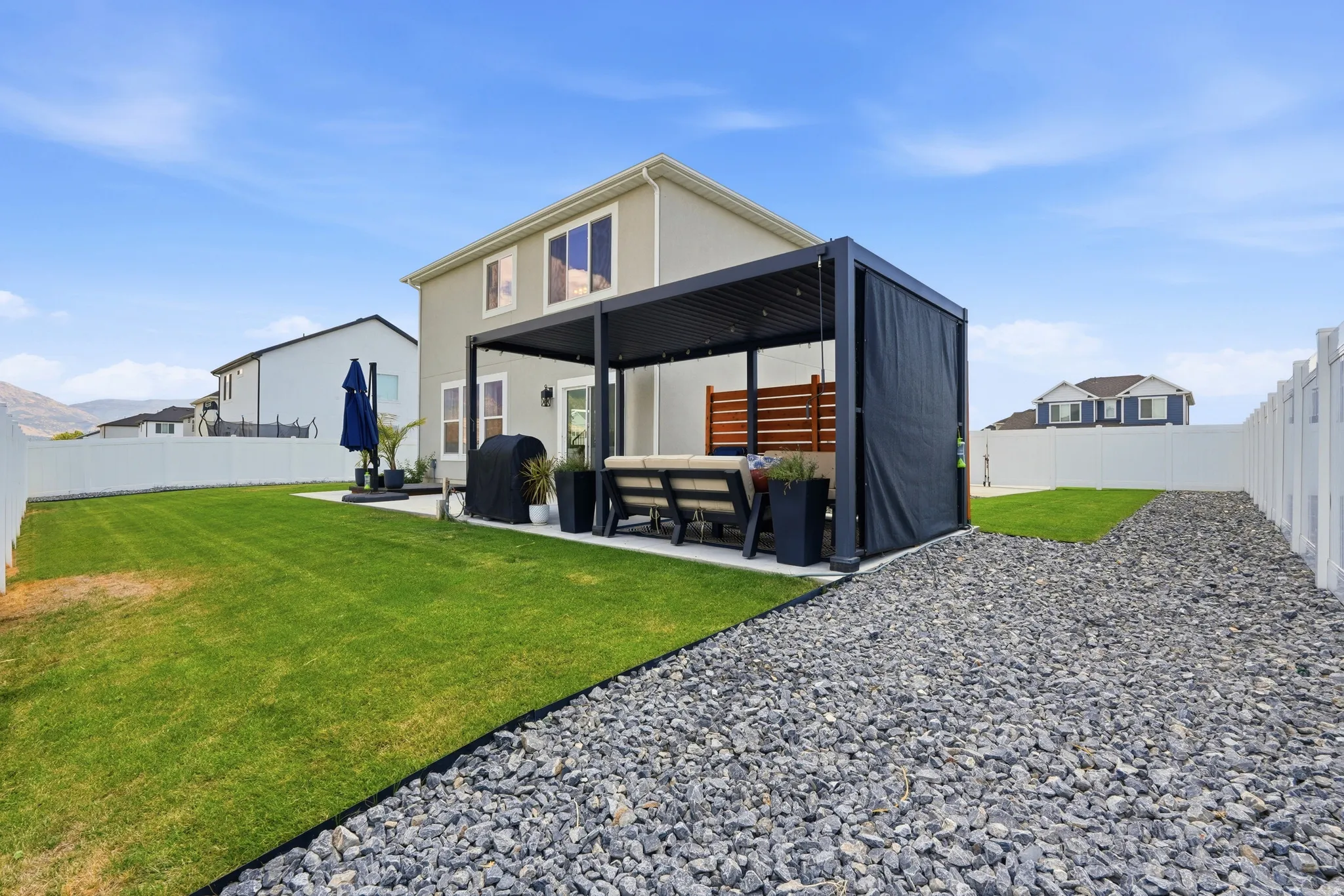 Expansive backyard featuring a lush lawn, white vinyl fencing, a concrete patio, a modern pergola with retractable shades, and a decorative gravel pathway