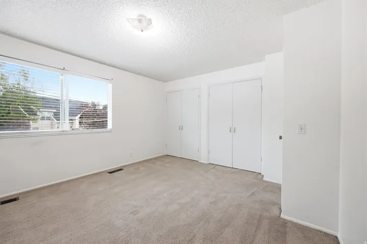 Neutral-toned carpeting spans this bright room, featuring a large window with horizontal blinds