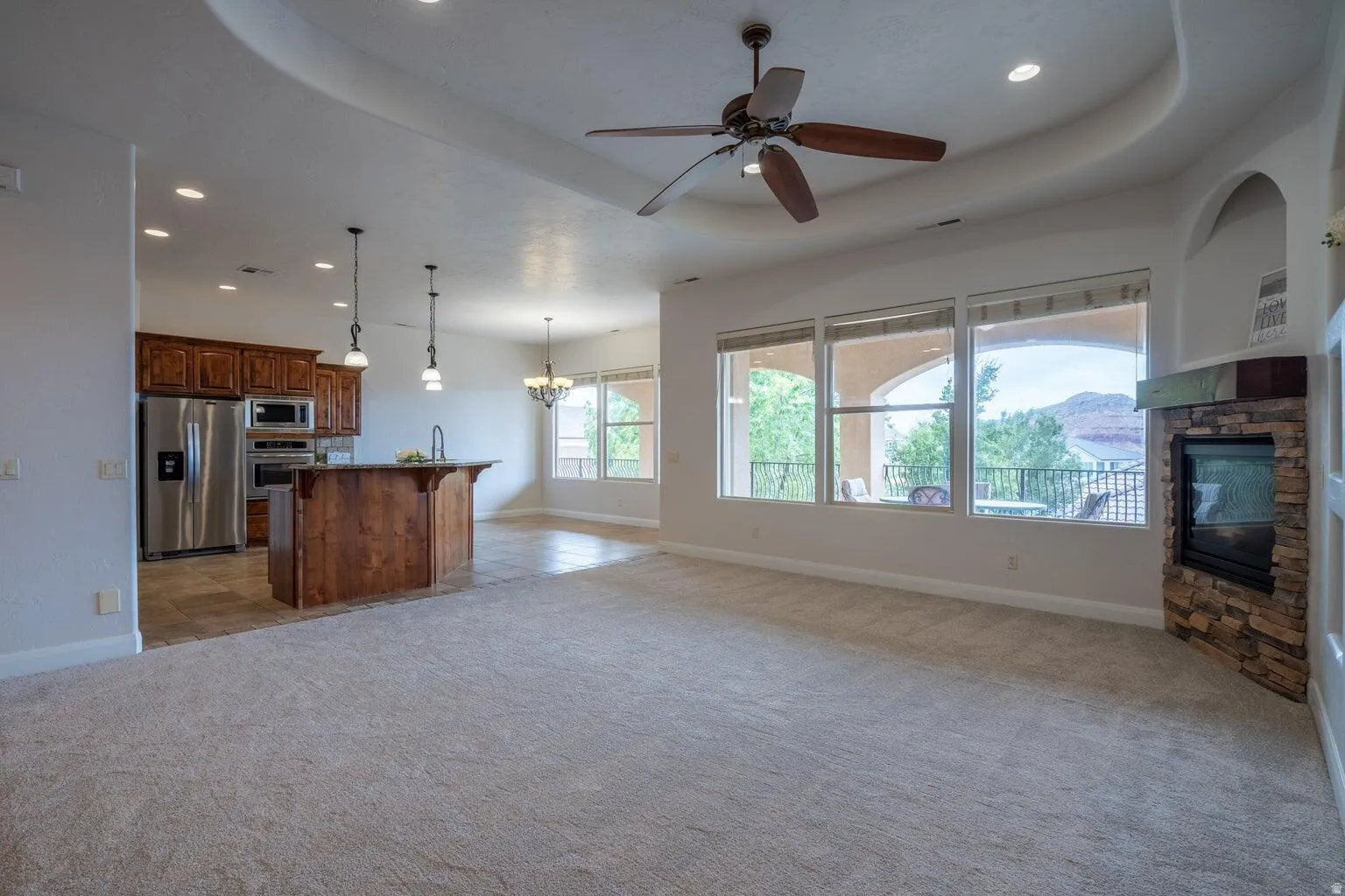 Open-concept living area featuring a stone-faced fireplace, a multi-pane window wall, and a tray ceiling with recessed lighting