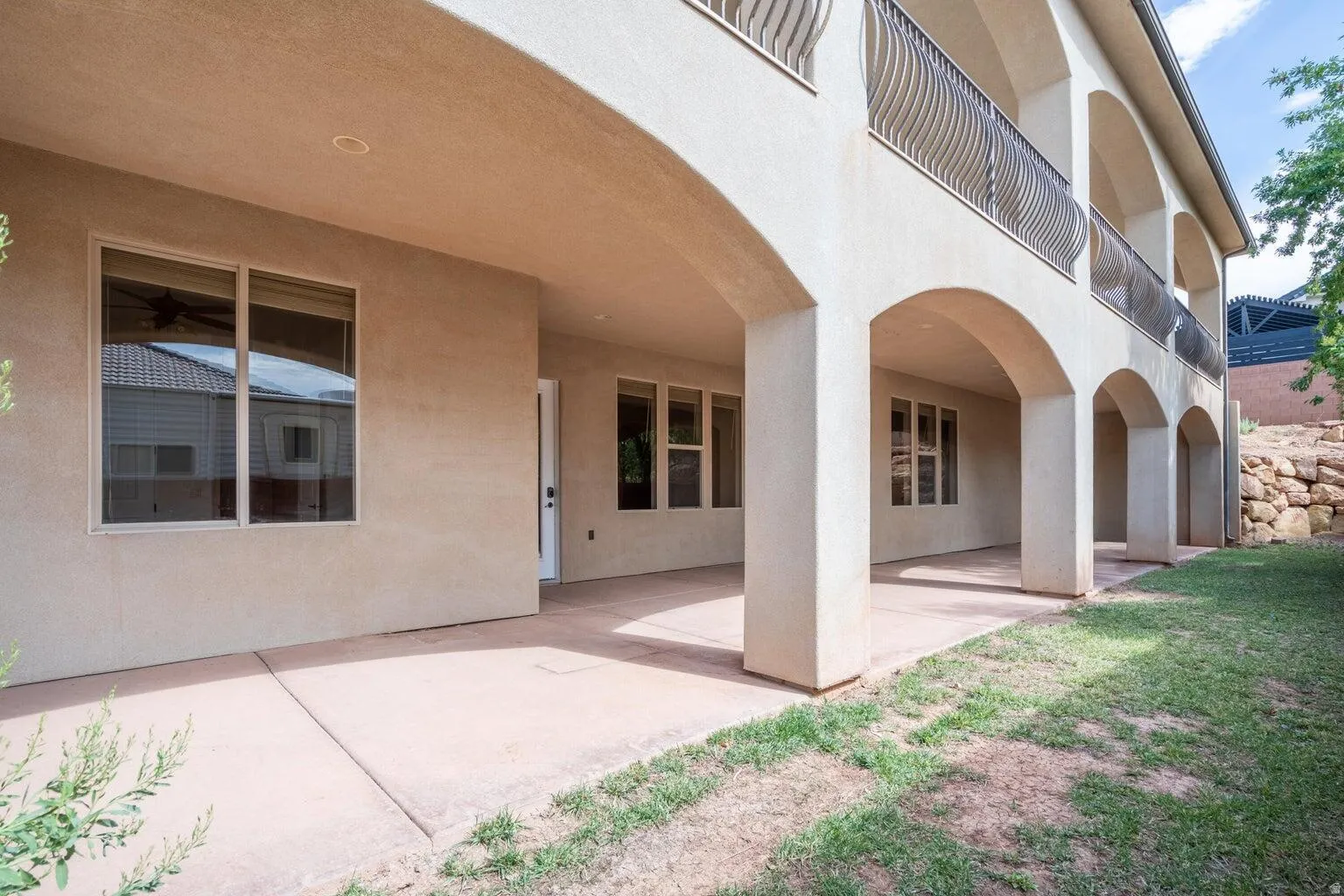 Covered patio with arched supports, stucco exterior, multiple large windows, and a second-story balcony with metal railings