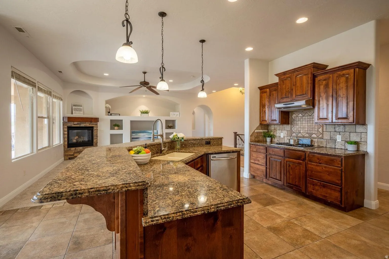 Open-concept kitchen and living area featuring granite countertops, wood cabinetry, tile flooring, a built-in fireplace with stone surround, and recessed and pendant lighting