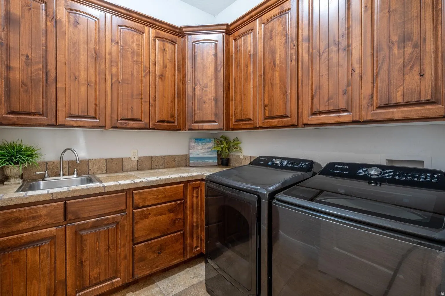 Functional laundry space featuring extensive wood cabinetry, a built-in sink with a gooseneck faucet, tiled countertops, and a complementary tiled backsplash